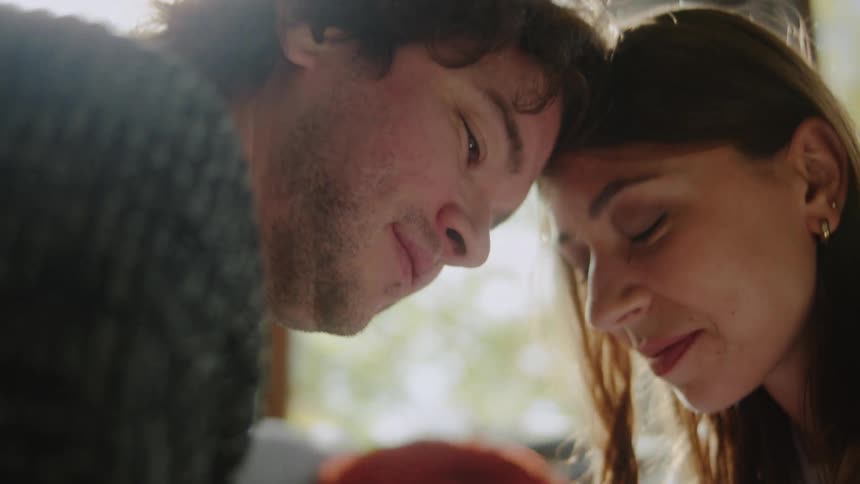 A couple bump their heads together in a romantic interaction as sunlight streams through the glass window into the room