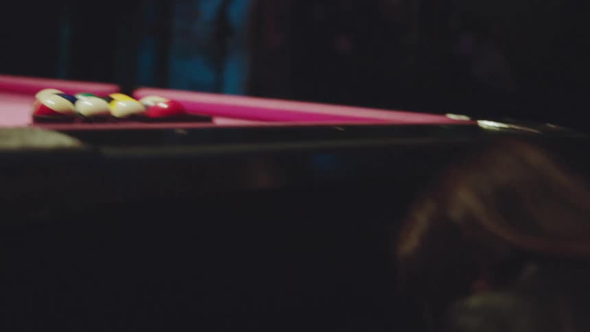 A woman lines up billiard balls on a brightly colored pink felt pool table under dramatic low light in a dimly lit club