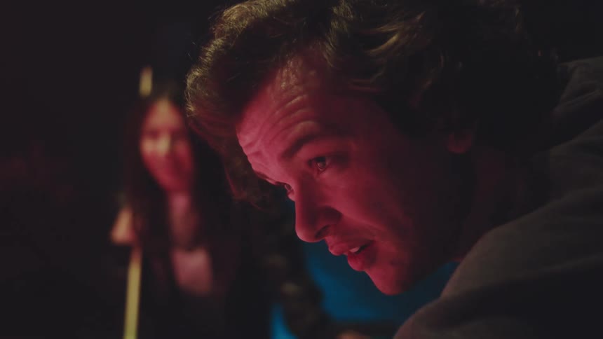 Young adult man leaning over a pool table to shoot a ball next to a young adult woman holding a cue next to him while both laugh and cheer indoors during night time