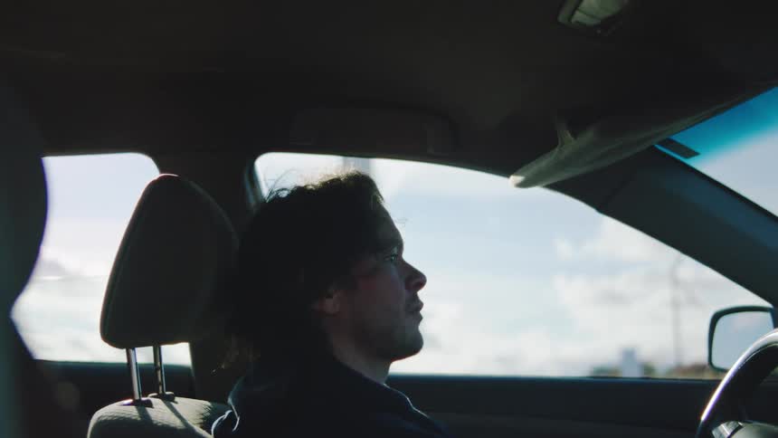 A man with short hair sitting in a car angrily punches the steering wheel and theres a wind turbine outside and a cloudy sky