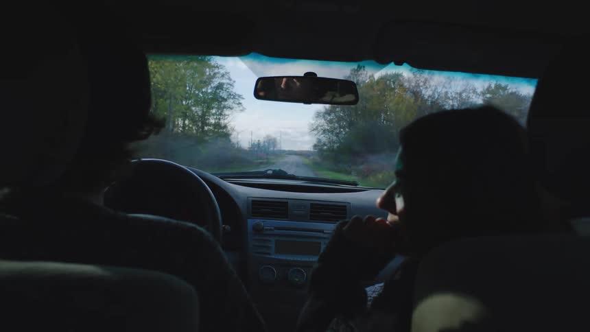 A man and woman drive down a tree lined road while having a conversation with one another