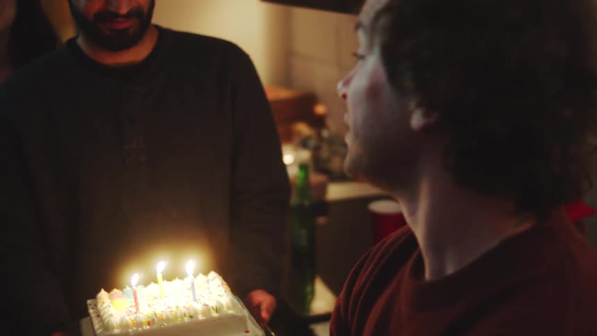 A man with curly hair smiles while talking then looks down to out candles on a birthday cake brought by a friend other friends gather around to celebrate