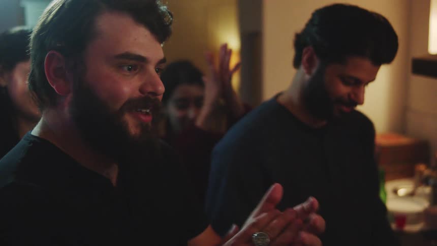 A young man is celebrated by friends and family on his birthday as a cake is brought to him he blows out the candles while everyone claps and afterward one of the men talks to him