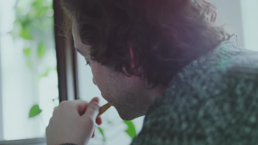 A young adult wearing a patterned sweater wets a wooden toothbrush under a running faucet in a welllit bathroom then begins brushing their teeth illuminated by bright daylight coming through a nearby window with an arm of a second person next to him