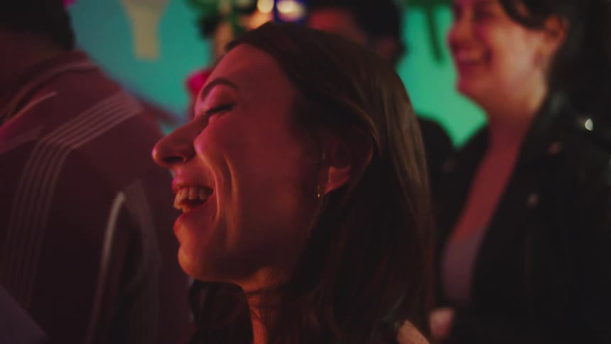 Young adult woman at a birthday party surrounded by other young adult people delivering an envelope and smiling indoors during night time