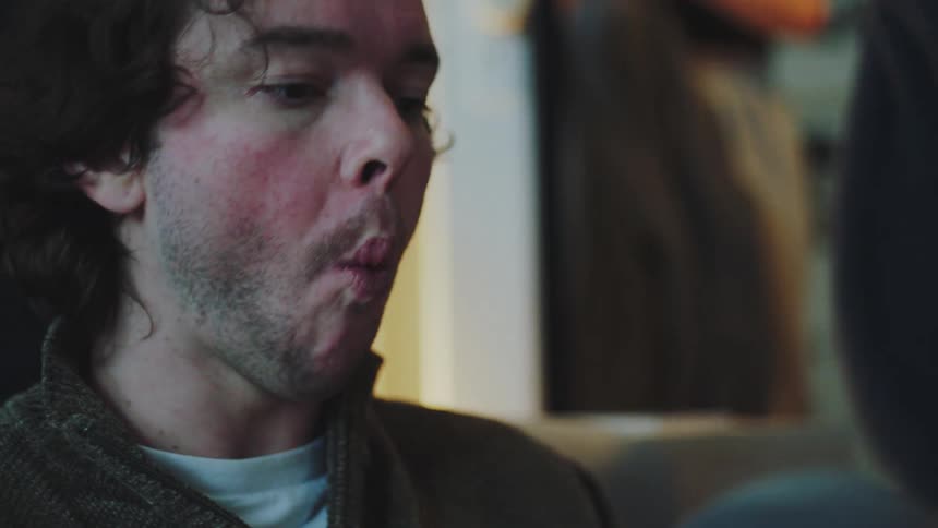 A young man takes a bite from a sandwich while sitting on a couch with a young woman behind him standing in front of a mirror