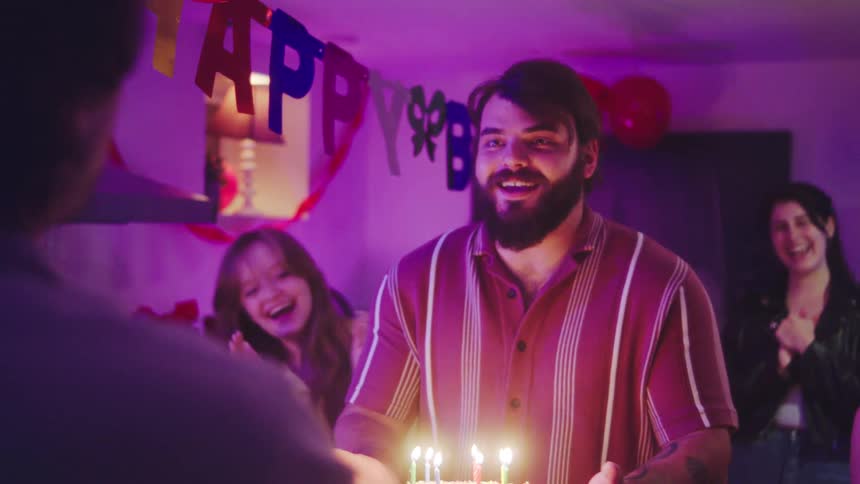 A person walks through a blue tinsel doorway to be greeted by a group of smiling friends holding a brightly sprinkled birthday cake with lit candles celebrating indoors under festive colorchanging party lights