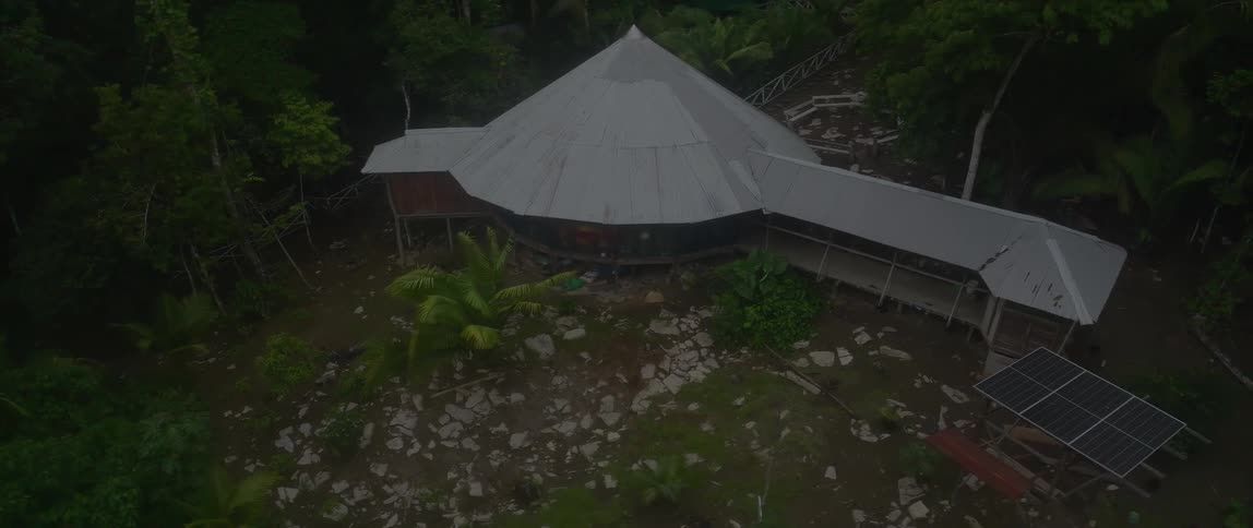 An aerial view shows a large circular building with a conical metal roof nestled within a dense tropical foresta solar panel is visible on the ground near the structure which is connected to a smaller outbuilding by a covered walkway