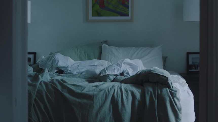 An unmade bed with rumpled sheets and pillows is viewed through a doorway in a dimly lit bedroom with a brightly colored piece of wall art hanging above it