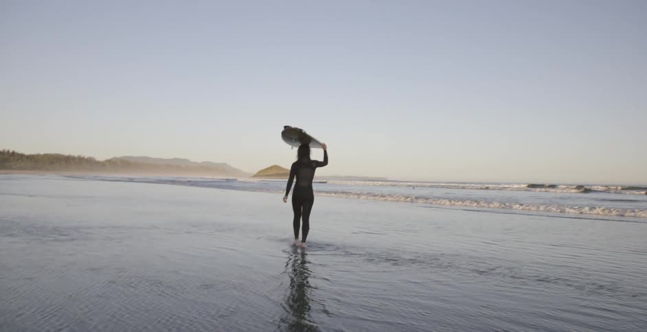 A woman walks with a surfboard on her head on a beach