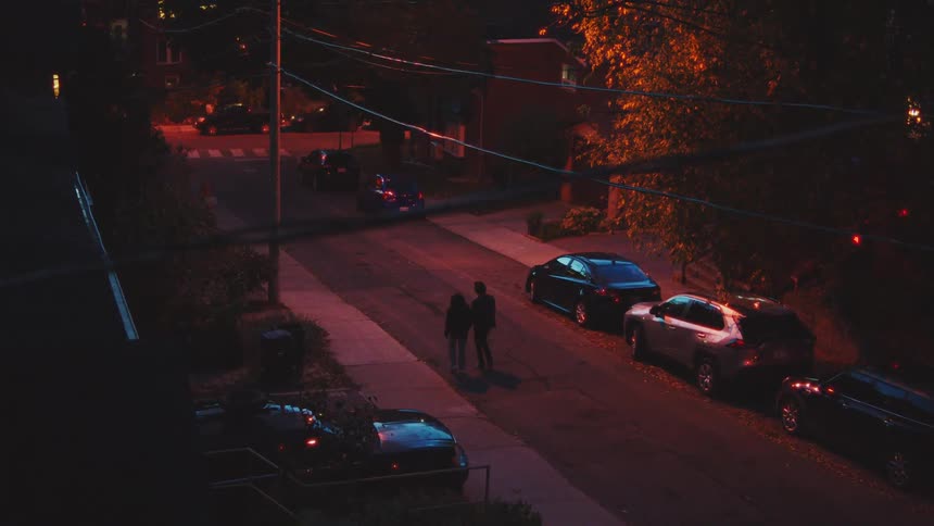 A young man and woman are walking together on a dimly lit street at night with parked cars and orange streetlights in a residential area