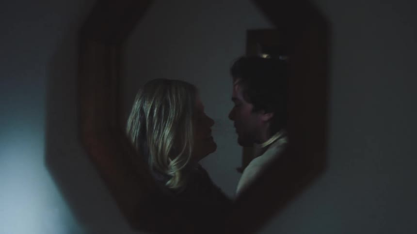 A man and a woman hug and laugh reflected in a dark octagonal mirror hanging in a dimly lit room