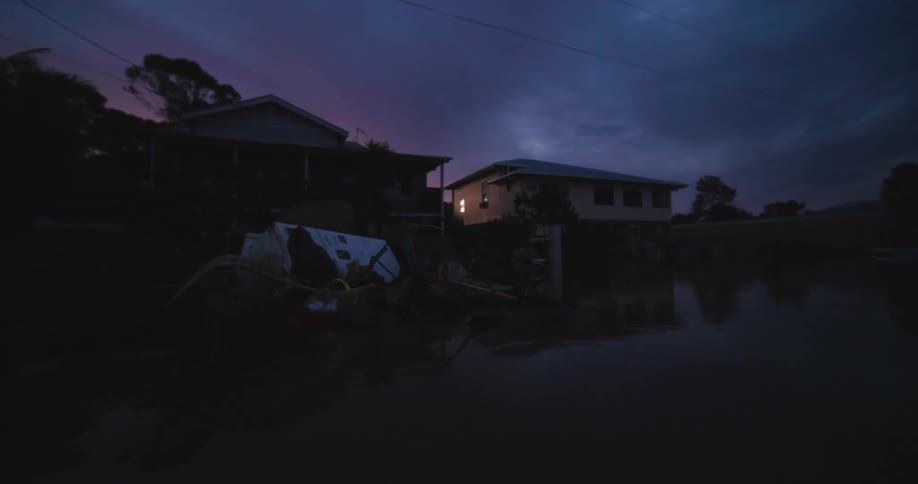 Two houses stand partially submerged in dark floodwater at night with debris floating nearby and a single window light shining under a deep purple and blue sky