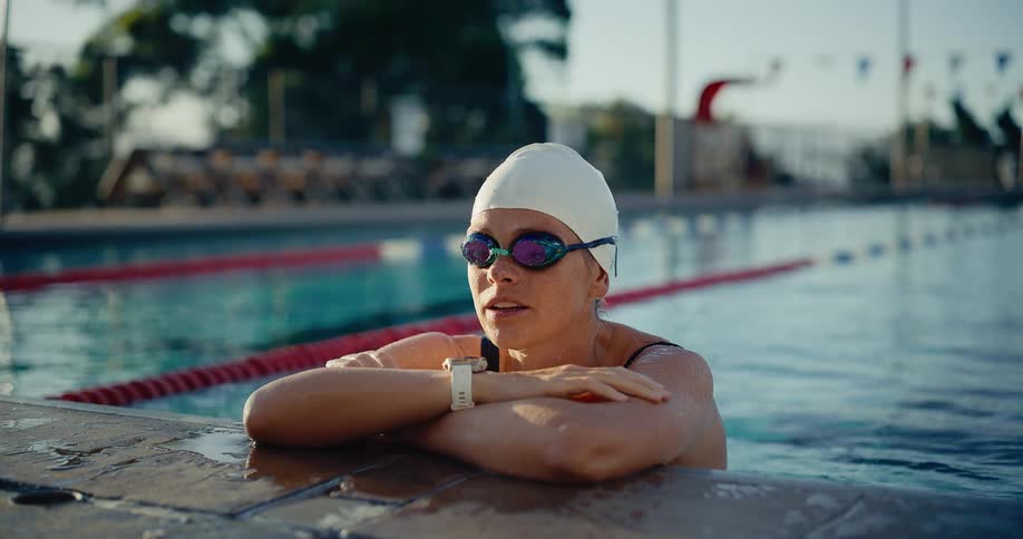 Swimmer in white cap and watch rests at pool edge beside flags and lounge chairs