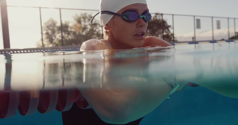 A woman with a white swim cap and mirrored green goggles sits at the edge of a pool breathing heavily after a swim