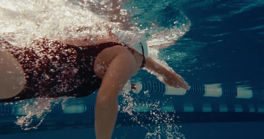 This video features an underwater lowangle shot of a swimmer in a black swimsuit white swim cap and goggles performing the freestyle strokethe surface of the water is brilliantly illuminated by sunlight creating a dynamic display of white foam an