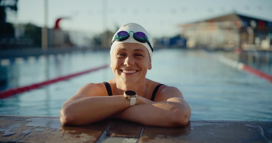 Woman wearing white cap and watch leaning on pool edge with lanes and structures in background
