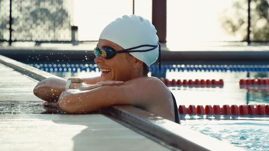 Young adult woman wearing a bathing suit and swim cap with goggles resting on the edge of a swimming pool and smiling during day time