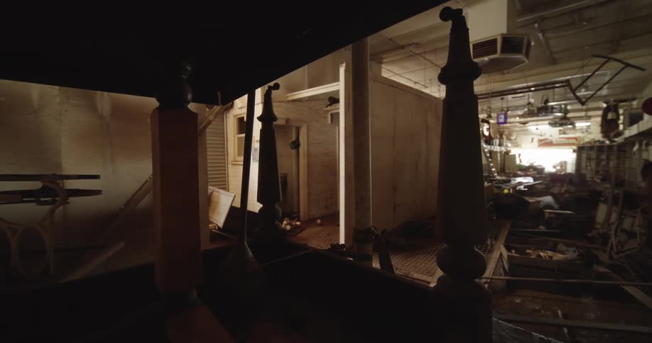 The heavily damaged and cluttered interior of a commercial building is viewed through the dark wooden frame of a four poster bed with bright light coming from the far end
