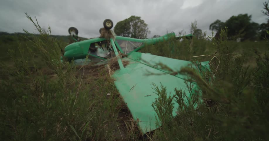 A small green ultralight aircraft lies overturned in a muddy field surrounded by tall green and brown brush under an overcast sky