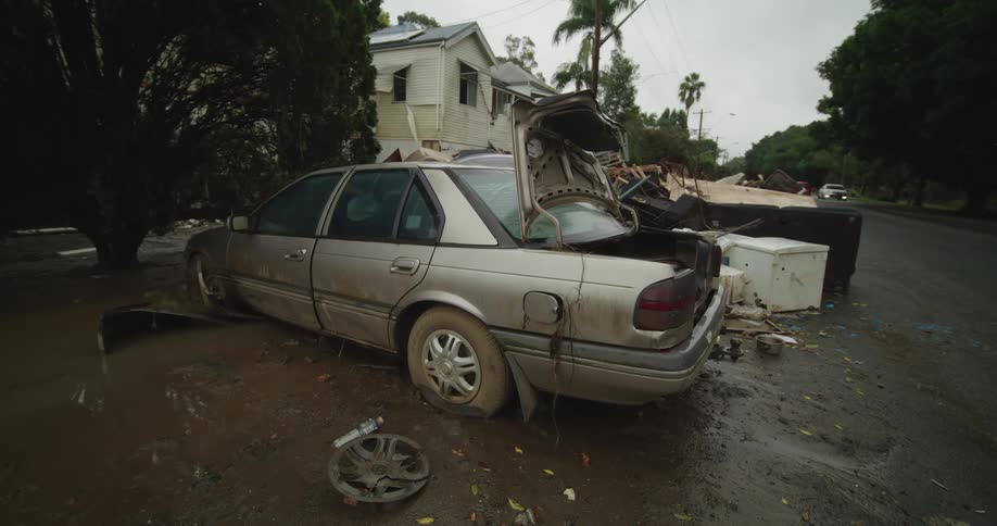 A dirty silver sedan sits abandoned on a muddy street its trunk and drivers side door ajar with debris scattered around it the scene depicts significant damage from a flood or natural disaster with a damaged house visible in the background