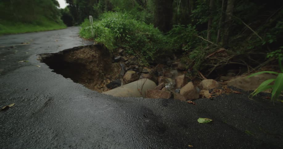 A wide shot captures a large section of an asphalt road in a dark wet forest that has completely washed away exposing a concrete culvert pipe and a pile of large rocks and debris below the gaping hole
