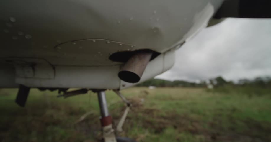 Exhaust pipe beneath aircraft fuselage rests on grassy field with cloudy sky in background