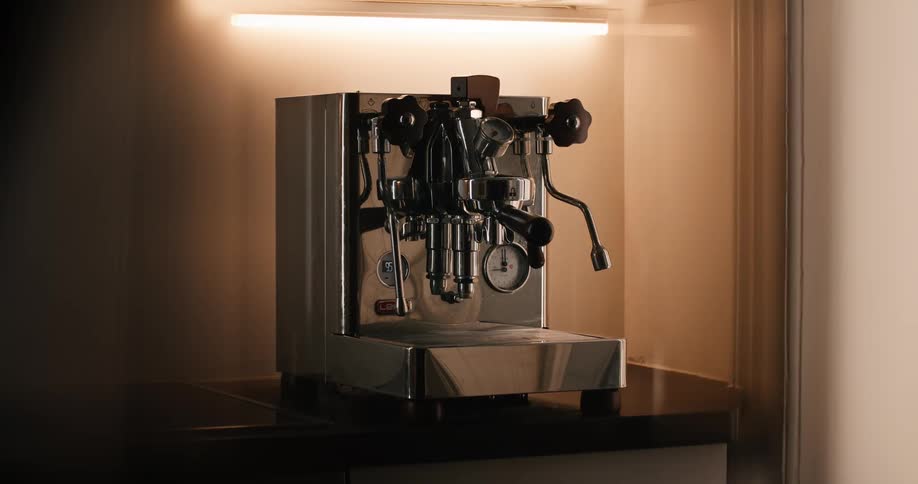 A chrome espresso machine on a dark counter is illuminated by a horizontal strip light that turns on above the machine