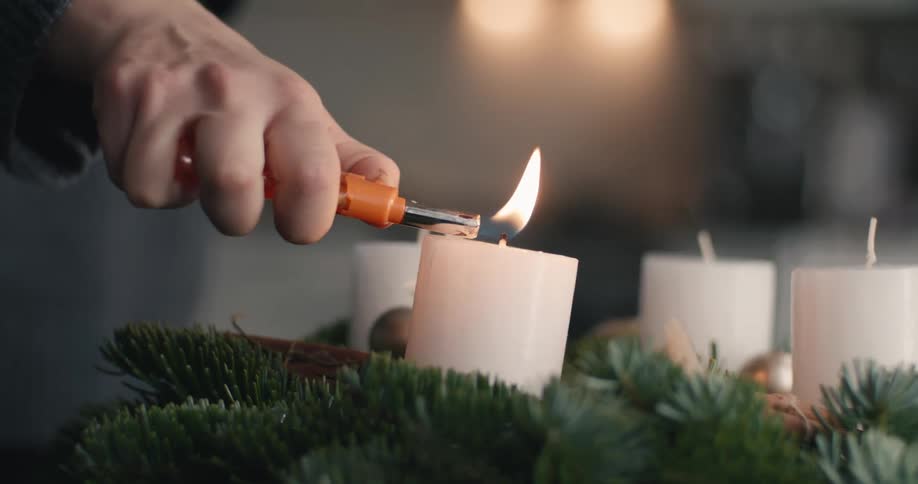 A close view of a womans hand lighting a white candle on a green pine advent wreath with an orange lighter