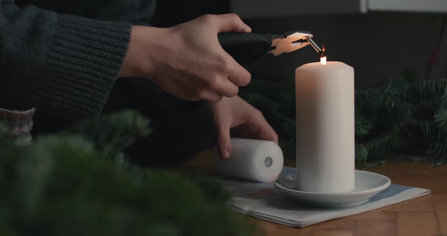 A person lights a large white pillar candle sitting on the floor amidst festive greenery