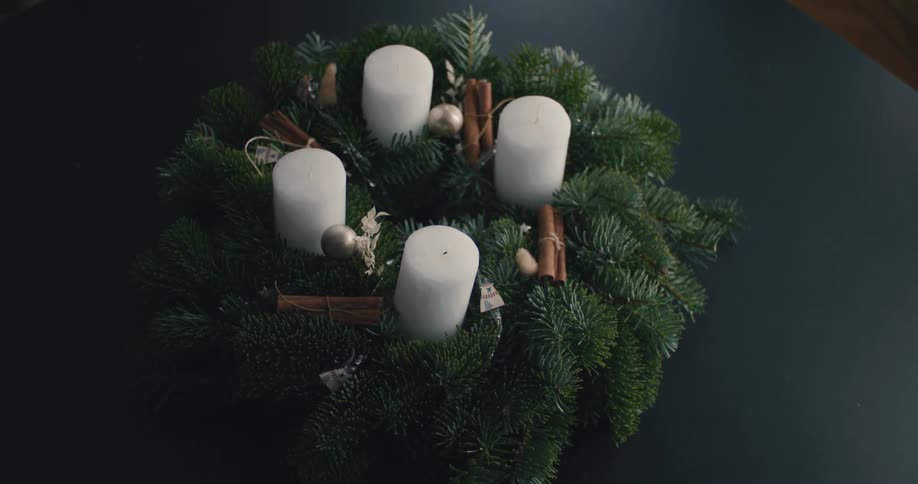 This video captures an advent wreath consisting of four white pillar candles nestled in a circular arrangement of evergreen boughsthe wreath is further decorated with cinnamon sticks and small silver or gold ornaments and a persons head is briefl