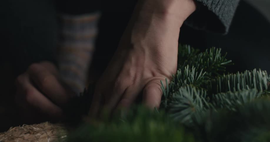 A closeup shot focuses on a persons hands wearing a gray sweater carefully manipulating fresh evergreen sprigs and wiring them onto a burlap base to construct a holiday wreath
