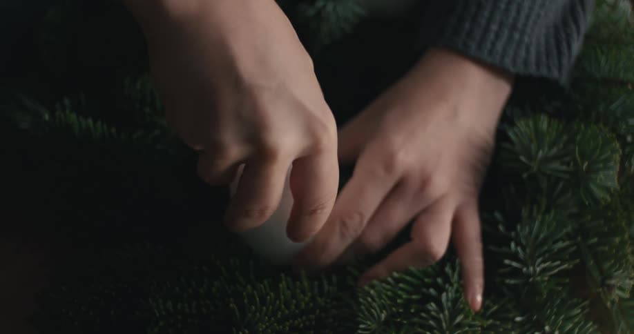 This video shows a closeup of a persons hands placing a white round pillar candle into an arrangement of dark green fir or pine needles suggesting the creation of an advent wreath or other seasonal decoration the final shot is a closeup of the perso