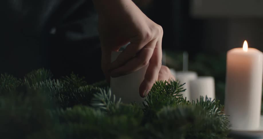A person carefully places a white candle into an evergreen wreath next to a burning candle