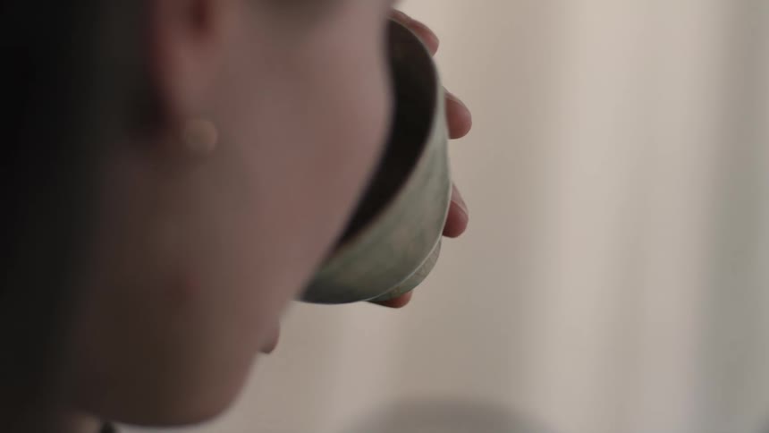 A woman raises a small textured light colored ceramic bowl to her mouth to drink in a dimly lit indoor space