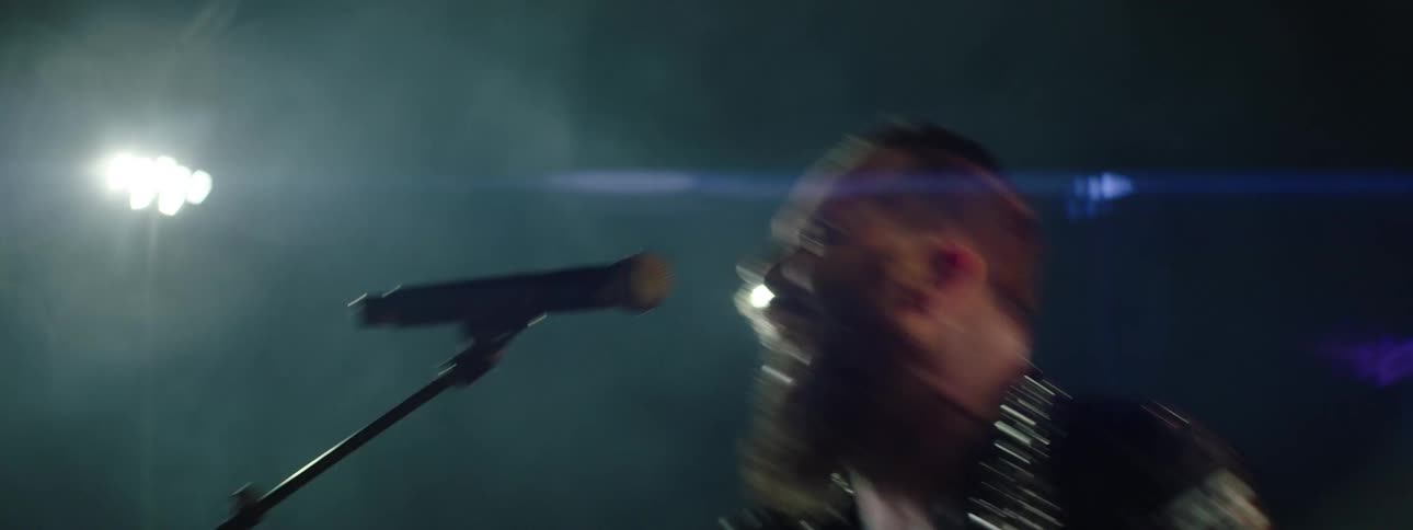 A close view shows a man with a full beard wearing a studded leather jacket singing aggressively into a microphone in a smoky bright stadium concert