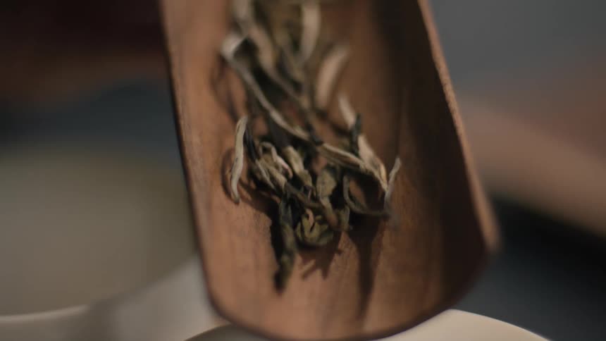 Wooden scoop tilted to pour dried tea leaves over white cup in blurred background