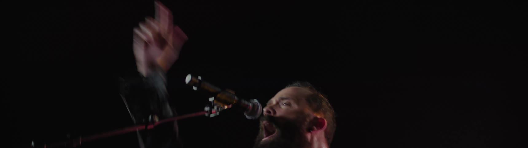 Bearded man singing on a microphone and playing an electric guitar next to another guitar player and a drummer on a rock concert at a stadium outdoors during night time