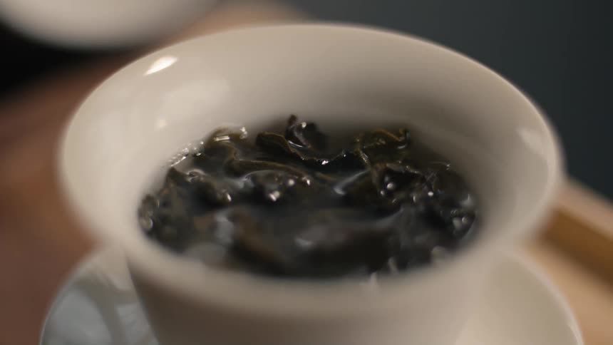 A closeup shot shows a hand removing the lid from a white porcelain teacup revealing dark wet tea leaves steeping in water steam gently rises from the cup as the leaves unfurl in the hot liquid