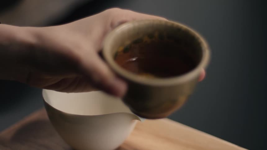 A hand holds a small earthytoned ceramic cup filled with a dark liquid possibly tea the hand then slowly lowers the cup over a larger white ceramic bowl suggesting the liquid is about to be poured or transferred