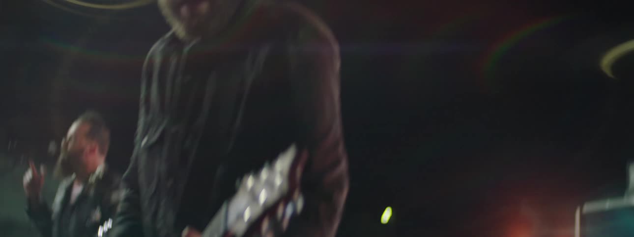 A male guitarist with a beard and dark clothing passionately plays an electric guitar on a brightly lit stage during a concert with a drummer visible behind him the video features dynamic camera movement lens flare and closeups capturing the high en