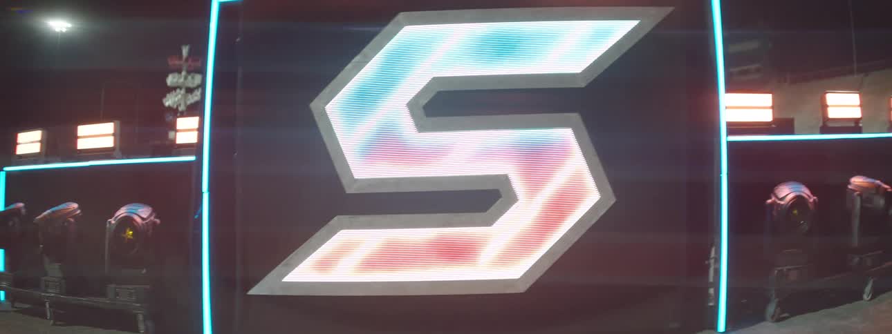 A closeup shot of a large led screen displays a stylized futuristic letter s logo with sharp edges glowing brightly with a shifting pattern of vibrant blue and red light effects