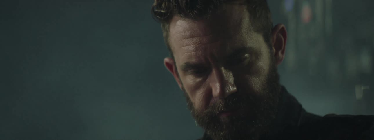 A man with a beard is concentrating while playing a striped electric guitar on a dark stage with mist and low light
