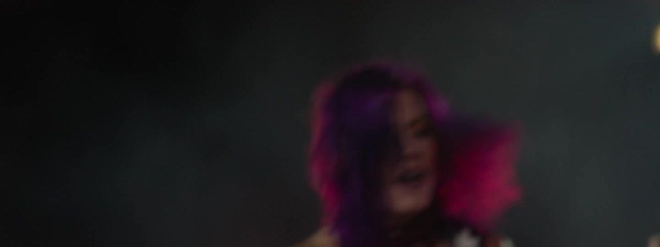 The video shows a closeup of a woman with vibrant purple hair dressed in a black top with a white lightning bolt graphic intensely playing a guitar on a dimly lit stage the camera movement is dynamic and shaky with flashes of red blue and white sta