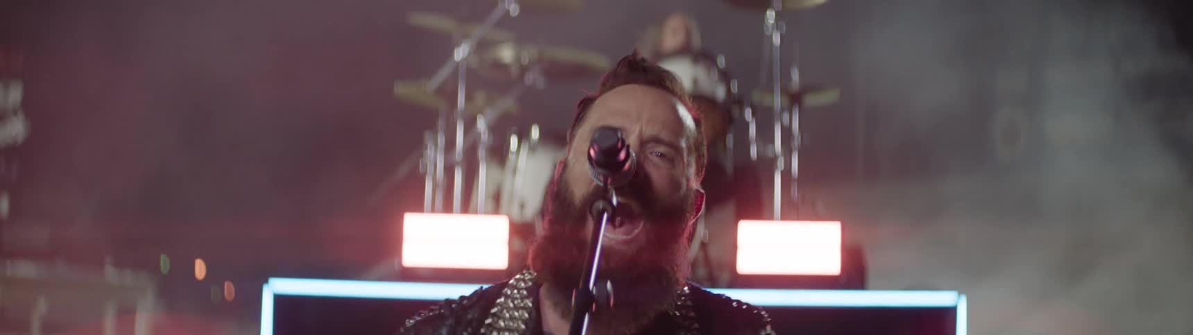 Bearded singer performs intensely in front of drums and rectangular lights in smoky setting