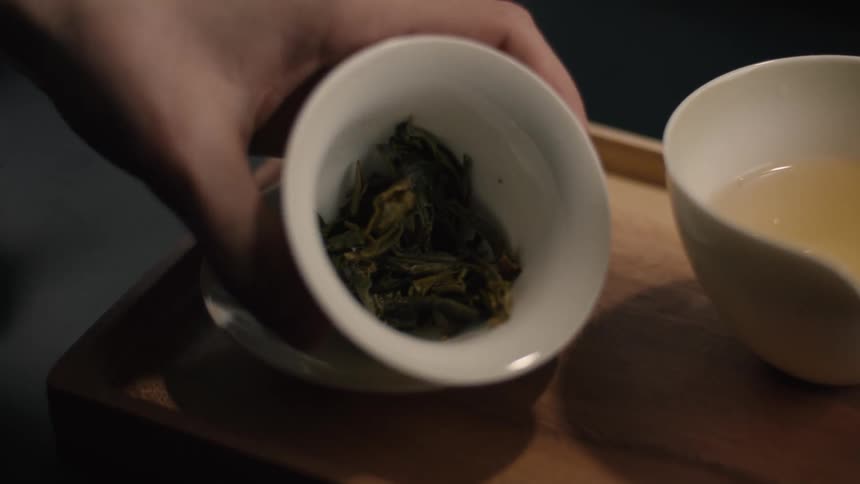 Hand holding vessel with tea leaves beside cup with light infusion on wooden tray