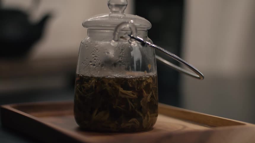 A glass teapot filled with hot water and loose green tea leaves steeps on a wooden tray in an indoor setting