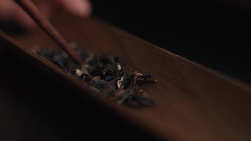 The video shows a closeup shot of dry tea leaves which appear to be dark and twisted resting in a long narrow curved wooden dish or traya persons hand uses a small wooden tool possibly a chopstick or specialized tea scoop to gently push and ar
