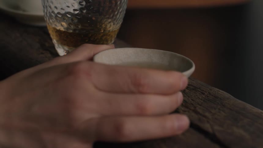 A persons hand picks up a cup of alcoholic drink with a glass beside it on a wooden table