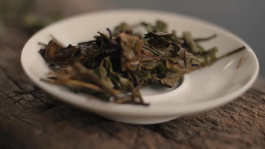 A close view shows a small white saucer holding dried brown and green tea leaves placed on a textured wooden table in a dimly lit indoor space